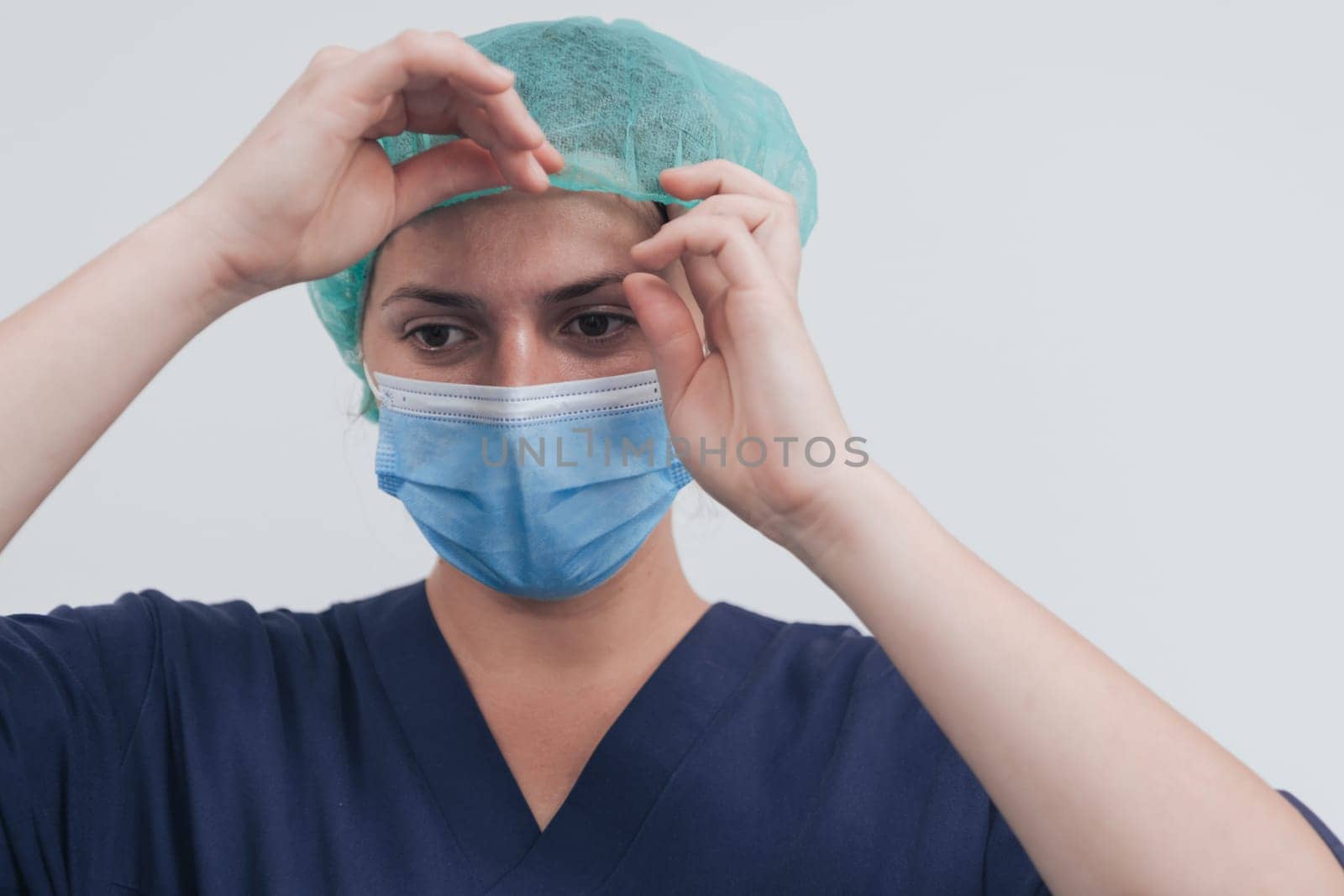 Close up of tired female doctor or scientist with a medical mask and surgical cap over grey background. Authentic real medical people hero in white in coronavirus pandemic is adjusting mask.