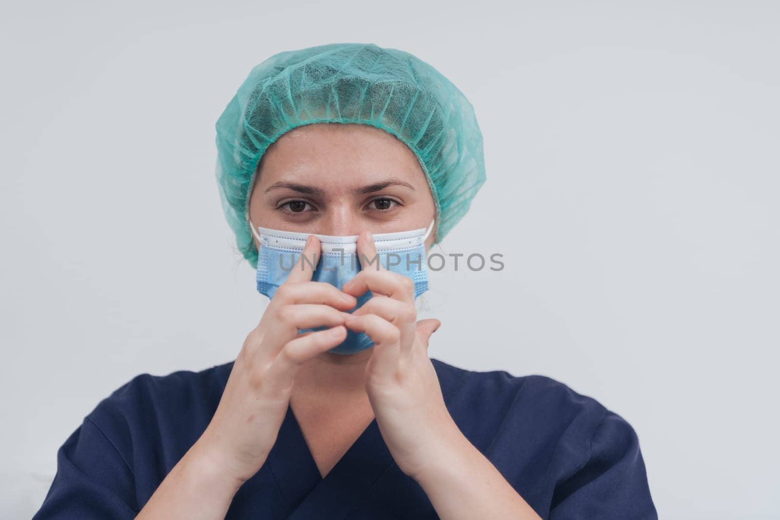 Close up of tired female doctor or scientist with a medical mask and surgical cap over grey background. Authentic real medical people hero in white in coronavirus pandemic is adjusting mask.