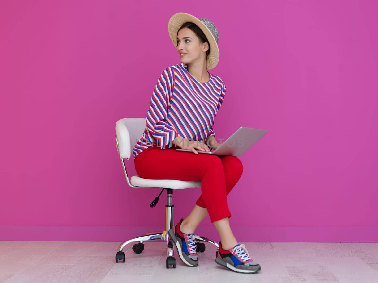 Portrait of young woman sitting on the chair and holding laptop on the lap isolated on pink background. Female model presenting fashion and technologz concept
