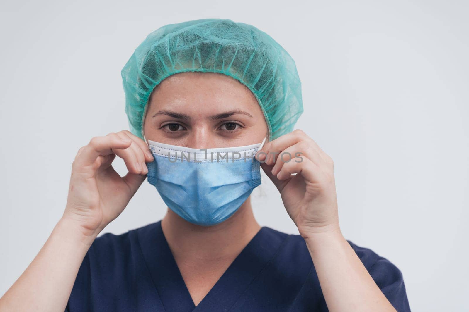 Close up of tired female doctor or scientist with a medical mask and surgical cap over grey background. Authentic real medical people hero in white in coronavirus pandemic is adjusting mask.