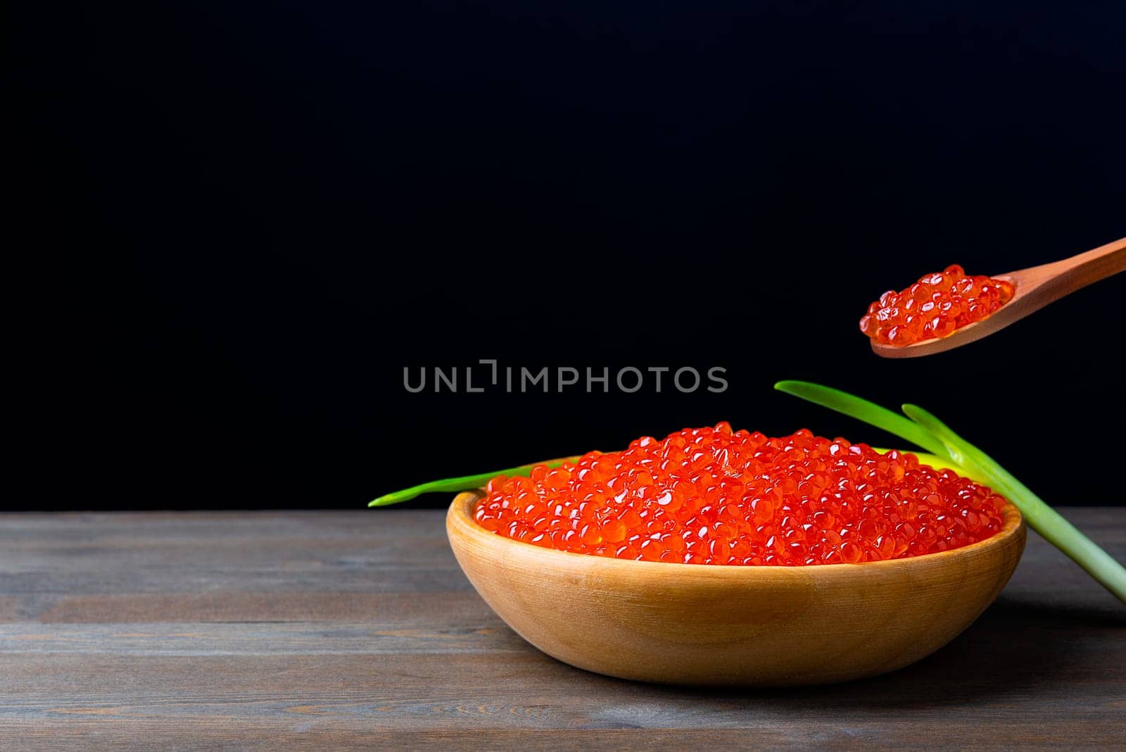 Red caviar in a wooden cup on a black background with a spoon and green onions. A large pile of bright caviar. Fresh delicious caviar. A place for advertising, logo, label, layout, mockup. Layout, template.