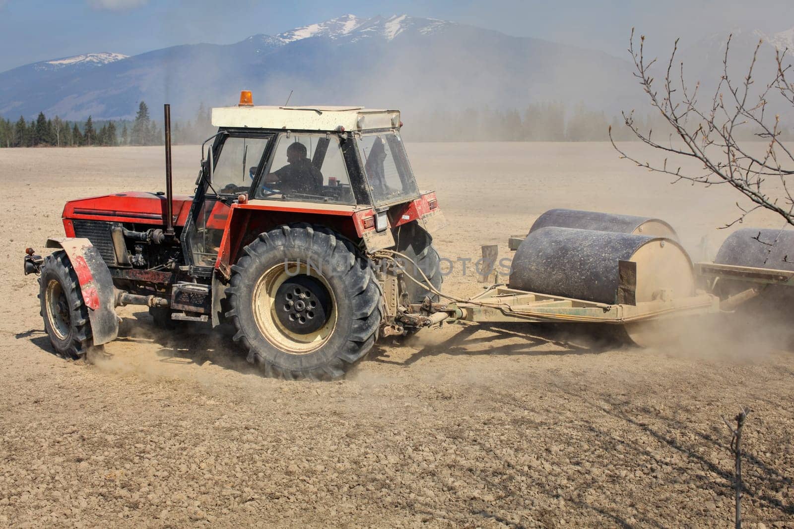 Liptovsky Hradok, Slovakia - April 22nd, 2018: Tractor turning when pulling heavy metal roller, preparing dry field in the spring, dust cloud behind, with mountains in the background.