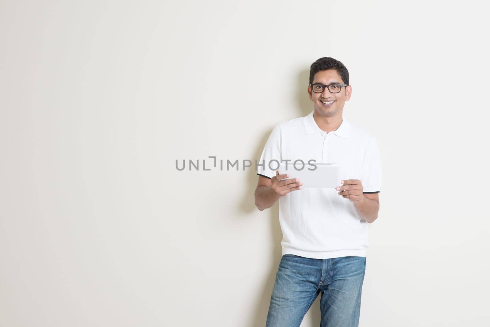Portrait of handsome Indian guy using tablet pc, standing on plain background with shadow, copy space at side.