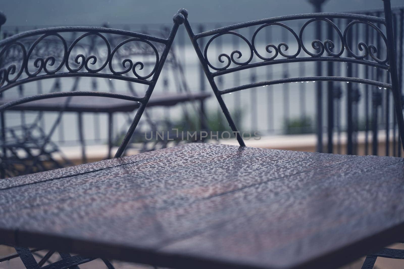 closeup detail of black iron metal chairs near wooden table with rain drops on rainy outdoor restaurant terrace