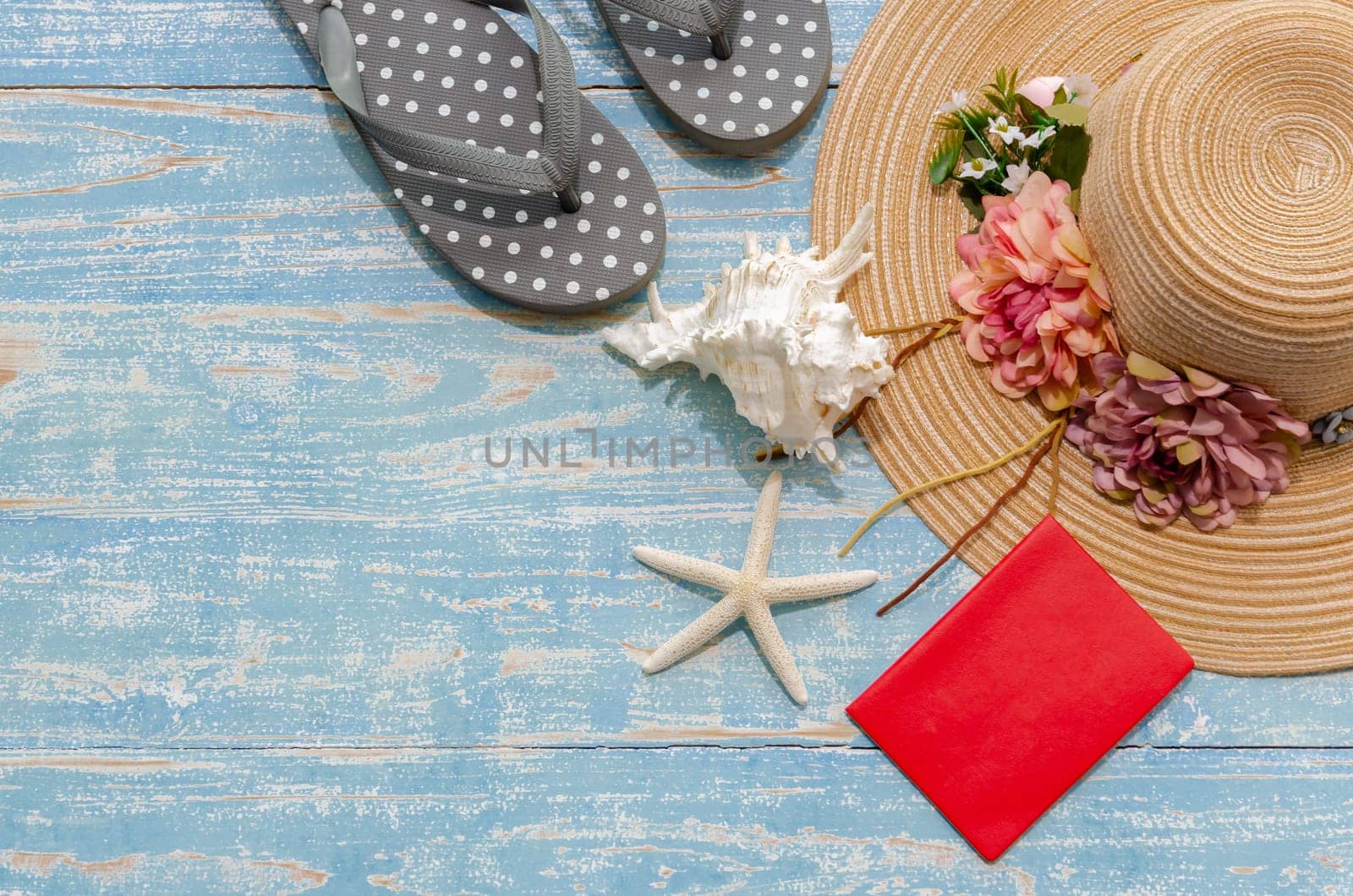 Flat lay summer beach accessories of lady flowered straw hat, flip flops and red passport with seashells and starfish on blue wooden background. Summer beach and holiday trip concept with copy space.