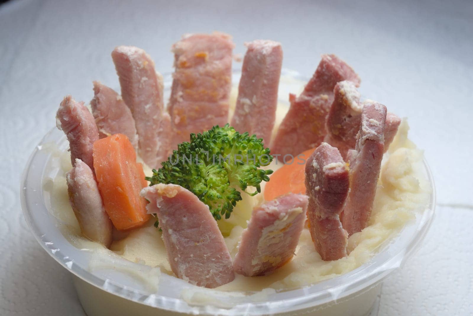Softly lit funny meal of gammon slices arranged in a circle, on top of a potato mash bed. In the middle is a broccoli floret and two carrot slices