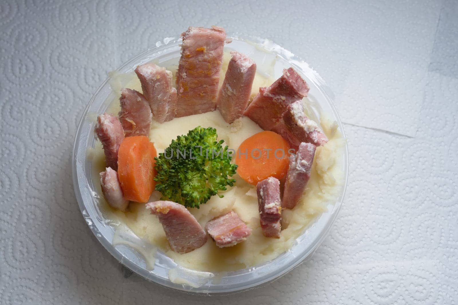 Tub of potato mash on top of white kitchen roll. Unusual presentation of meat and vegetables. Homemade twist on a supermarket ready meal