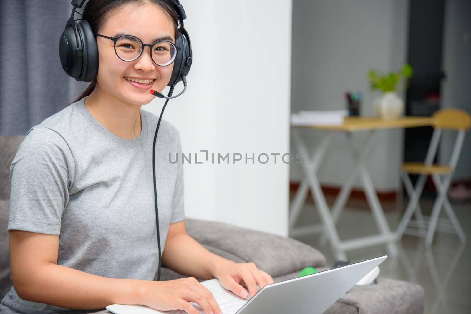 Asian young woman student with headphones sitting on the sofa smile looking up happy study online class college learning internet education, Teenage girl work distance on a laptop computer from home