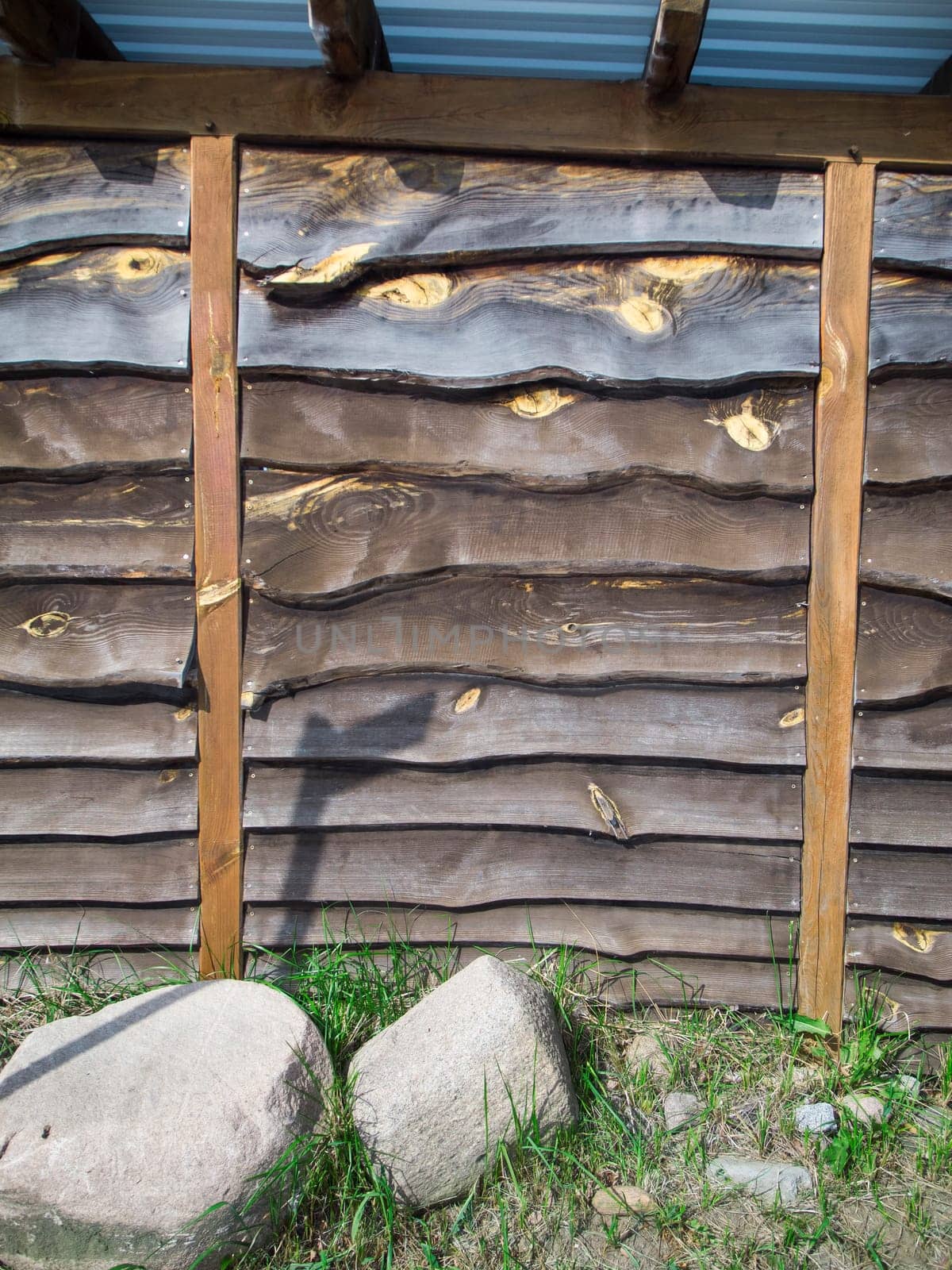 Format-filling view of a wall made of horizontally overlapping and untrimmed boards with two vertical pillars to stabilize the outside area and two field stones in front of it.