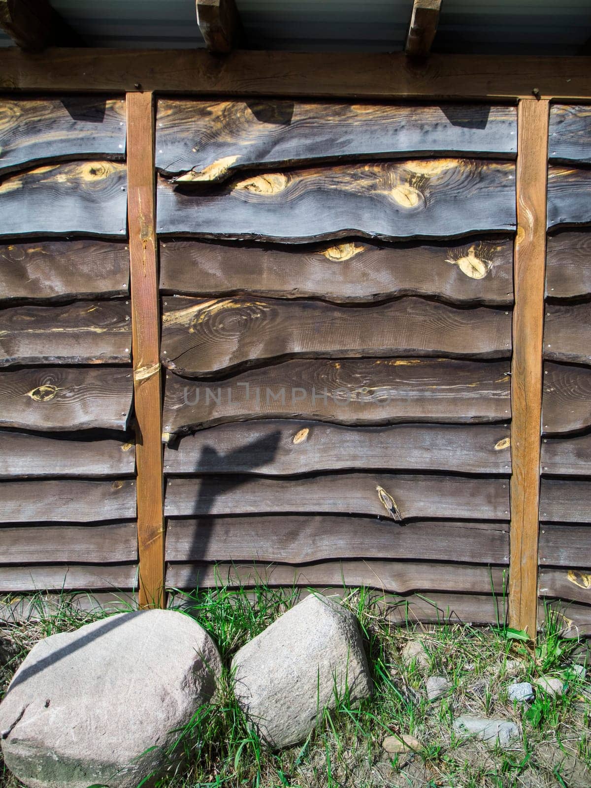 Format-filling view of a wall made of horizontally overlapping and untrimmed boards with two vertical pillars to stabilize the outside area and two field stones in front of it.