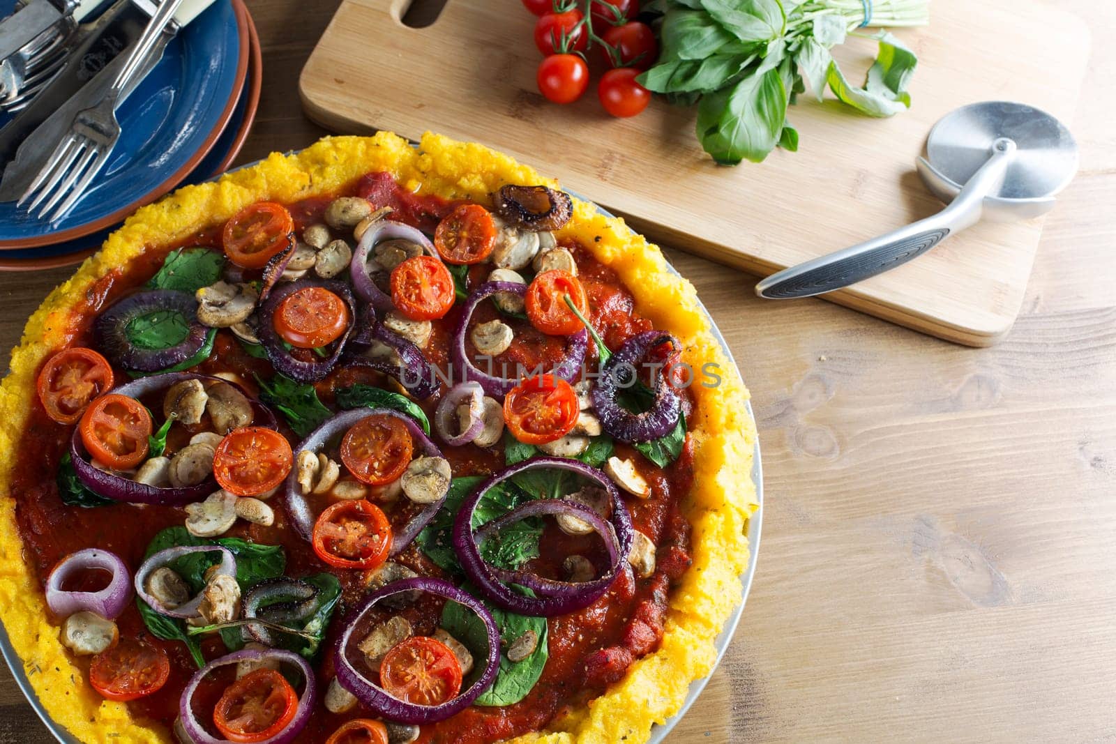 Whole vegan polenta pizza with cherry tomatoes, mushrooms, spinach and red onions.  Pizza slicer and ingredients in background with room for copy in lower right corner, horizontal orientation.