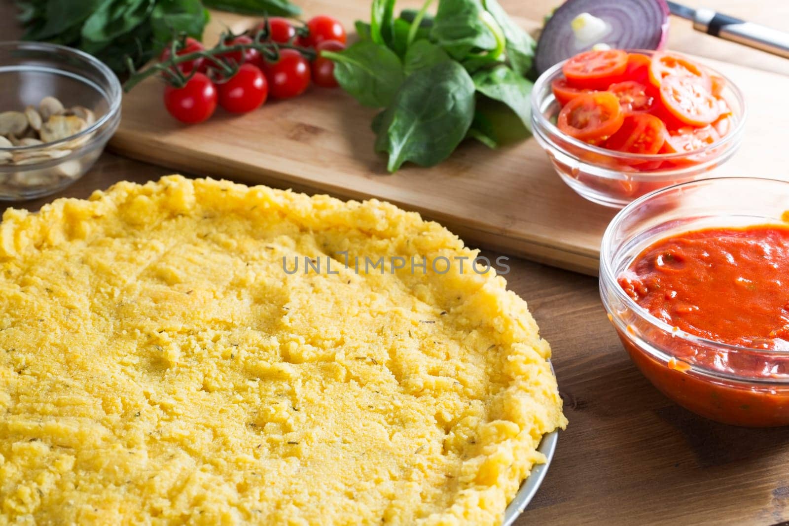Baking polenta pizza, polenta pizza crust ready for toppings with bowl of tomato sauce to the side and tomatoes, spinach and mushrooms next to pizza crust.