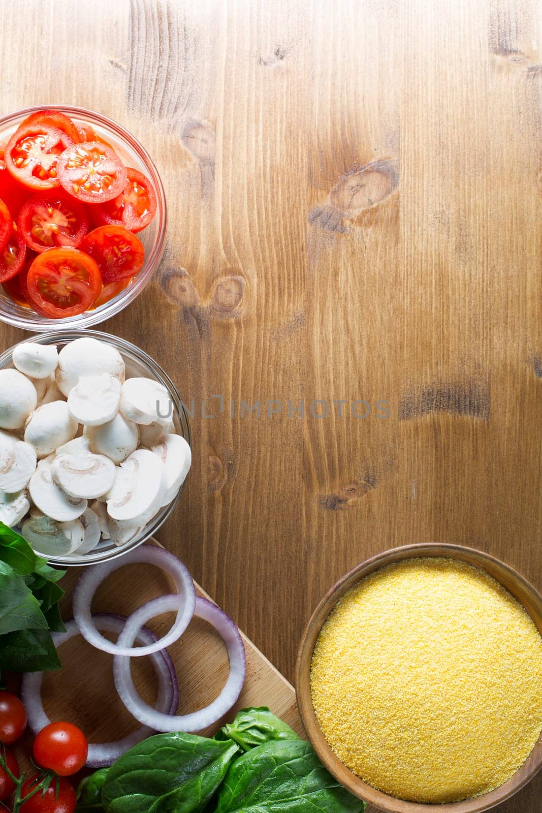 Ingredients for polenta recipe, with polenta in wooden bowl with sliced tomatoes, spinach and red onions.  Viewed from angle directly above with copy space on wooden table.  Can be used vertical or horizontal.