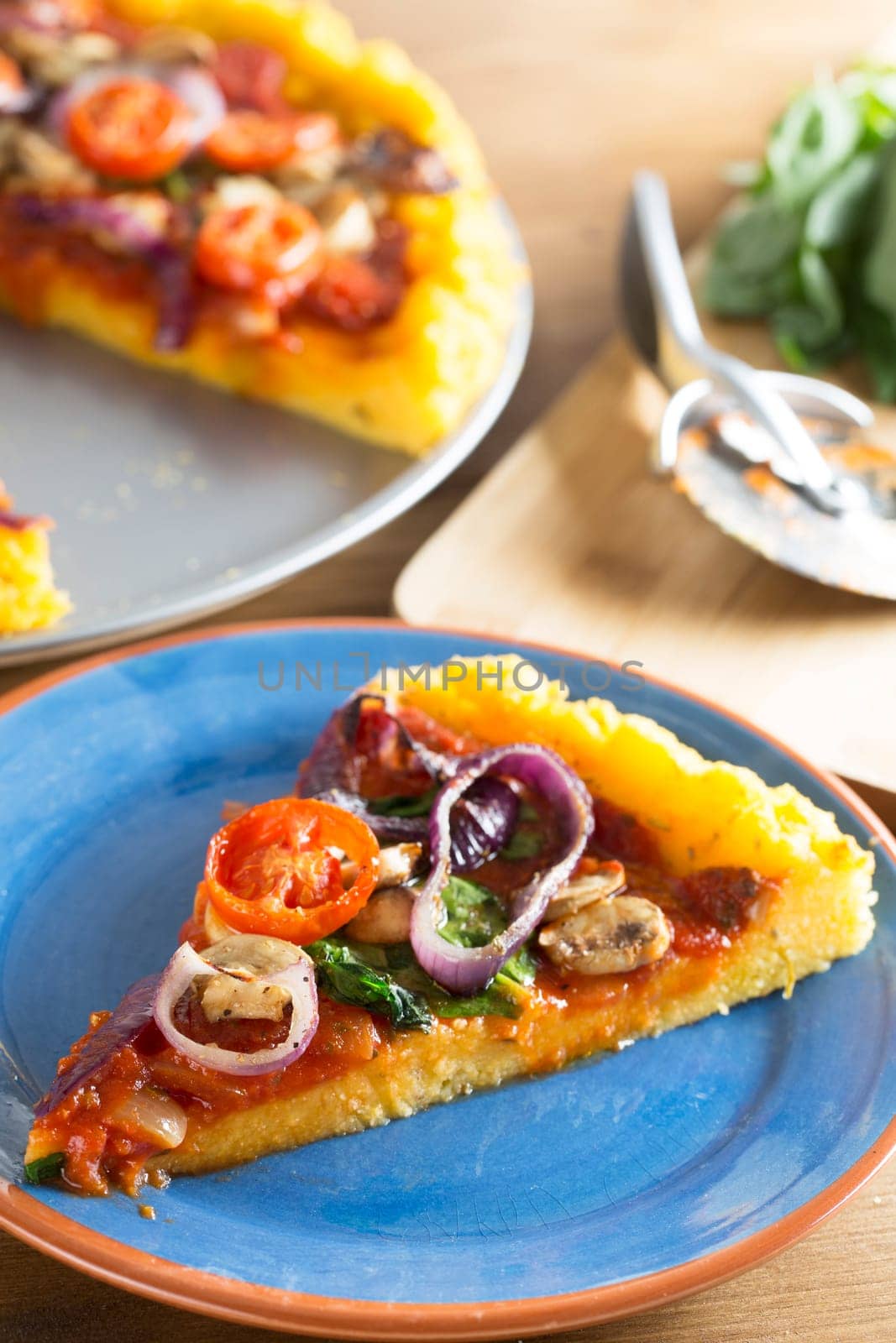 Vegan polenta pizza slice with cherry tomatoes, mushrooms, spinach and onion, on blue plate with whole pizza and ingredients out of focus in background giving room for copy.  Vertical orientation.