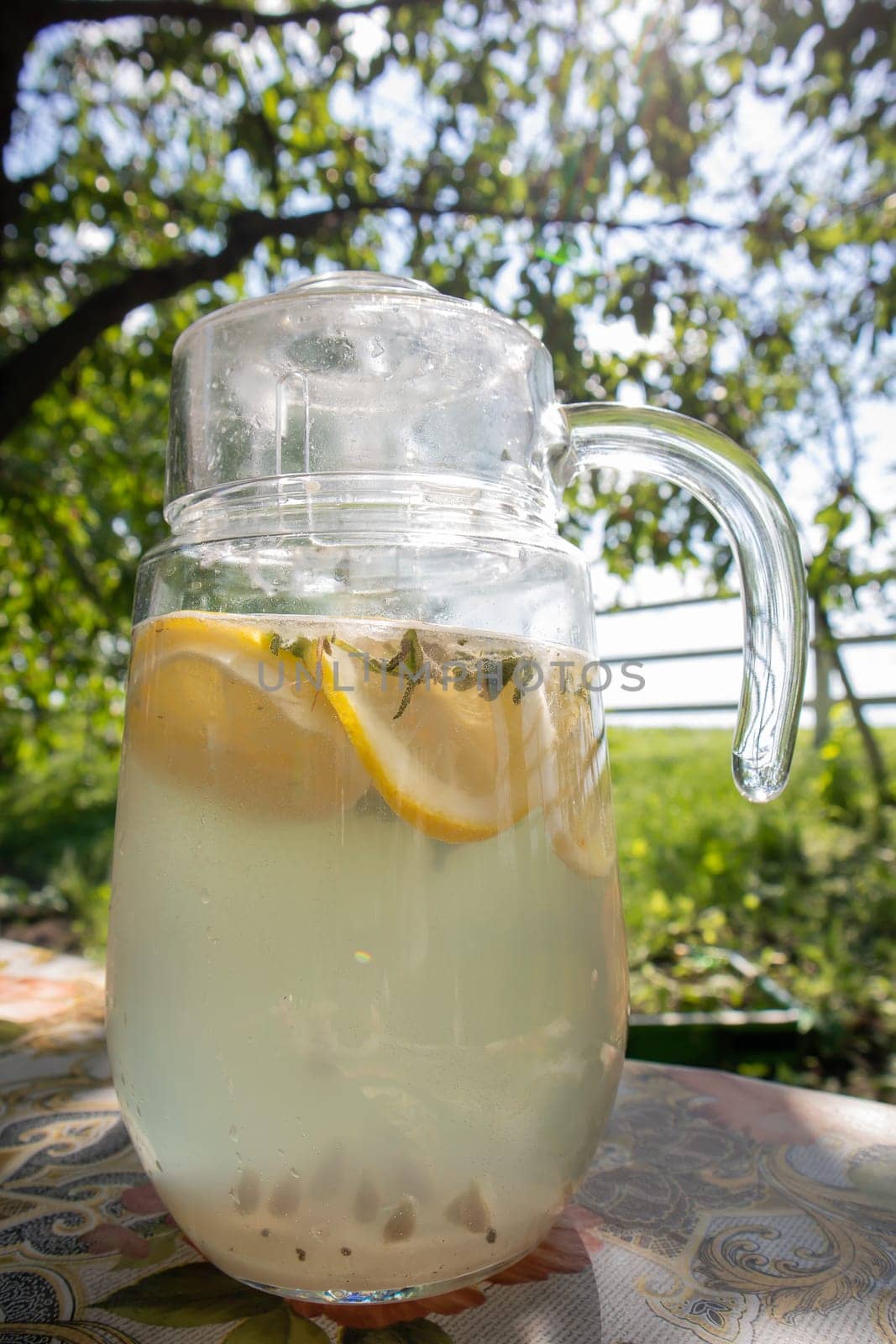 Homemade lemonade made from lemons in a large glass jug on the table in the garden. A jug with lemon and mint stands on the street against the backdrop of greenery on a hot summer day