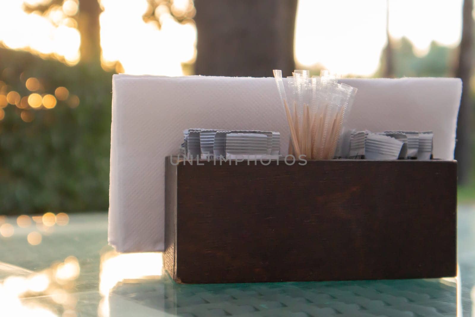 A brown wooden napkin holder and toothpicks sit on the glass table of the cafe, illuminated by the sun's rays. Table setting in the restaurant. Glass table in a green outdoor garden