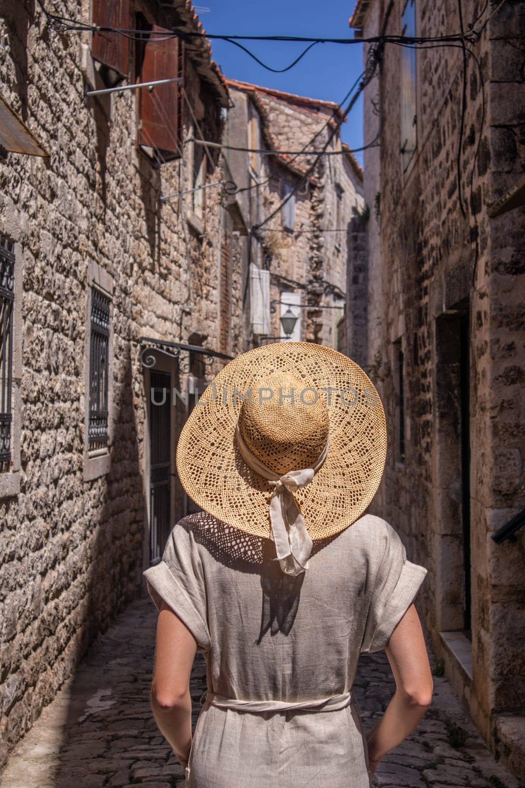 Beautiful blonde young female traveler wearing straw sun hat sightseeing and enjoying summer vacation in an old traditional costal town at Adriatic cost, Croatia. Rear view against the old stone town.