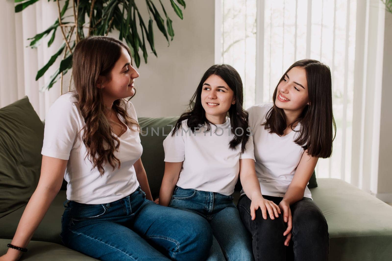 Three pretty smiling caucasian girls in white shirt sitting on the green sofa. Friends emotionally talking and discussing last news and life, sharing their thoughts and having some gossip topics