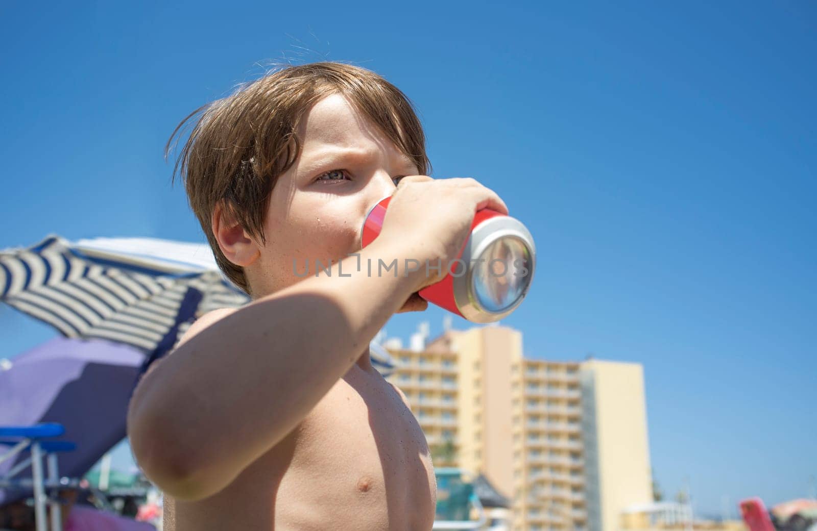 Little boy drinking from soft drink can. Beach background full of beach furniture