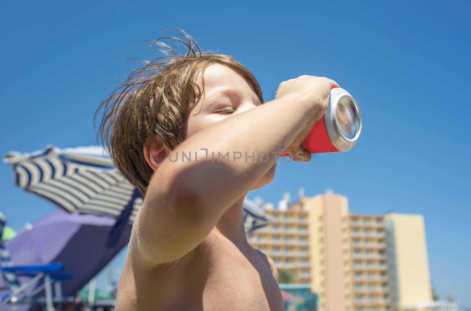 Little boy drinking from soft drink can. Beach background full of beach furniture