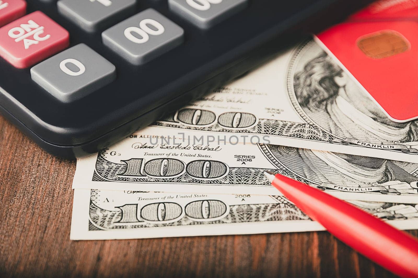 Dollars in one-hundred-dollar bills are spread out on the table next to a calculator, a credit card, and a pen. One-hundred-dollar bills. US dollars. Color style. Layout, template, mockup.