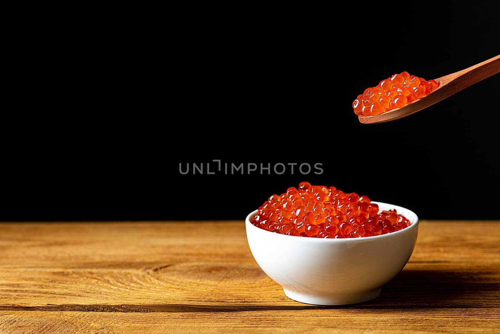 Red caviar in a wooden cup on a black background with a spoon. A large pile of bright caviar. Fresh delicious caviar. A place for advertising, logo, label, layout, mockup. Layout, template.