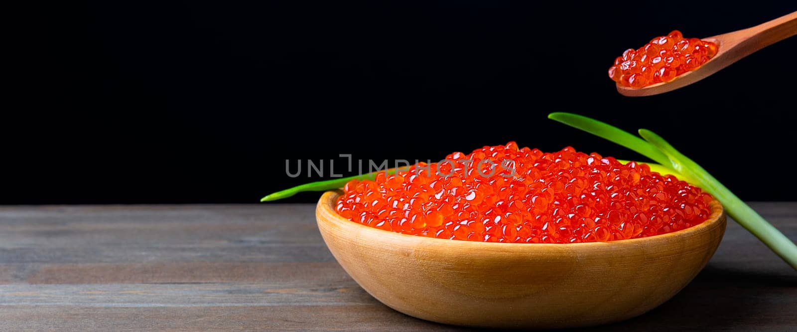 Red caviar in a wooden cup on a black background with a spoon and green onions. A large pile of bright caviar. Fresh delicious caviar. A place for advertising, logo, label, layout, mockup. Layout, template.