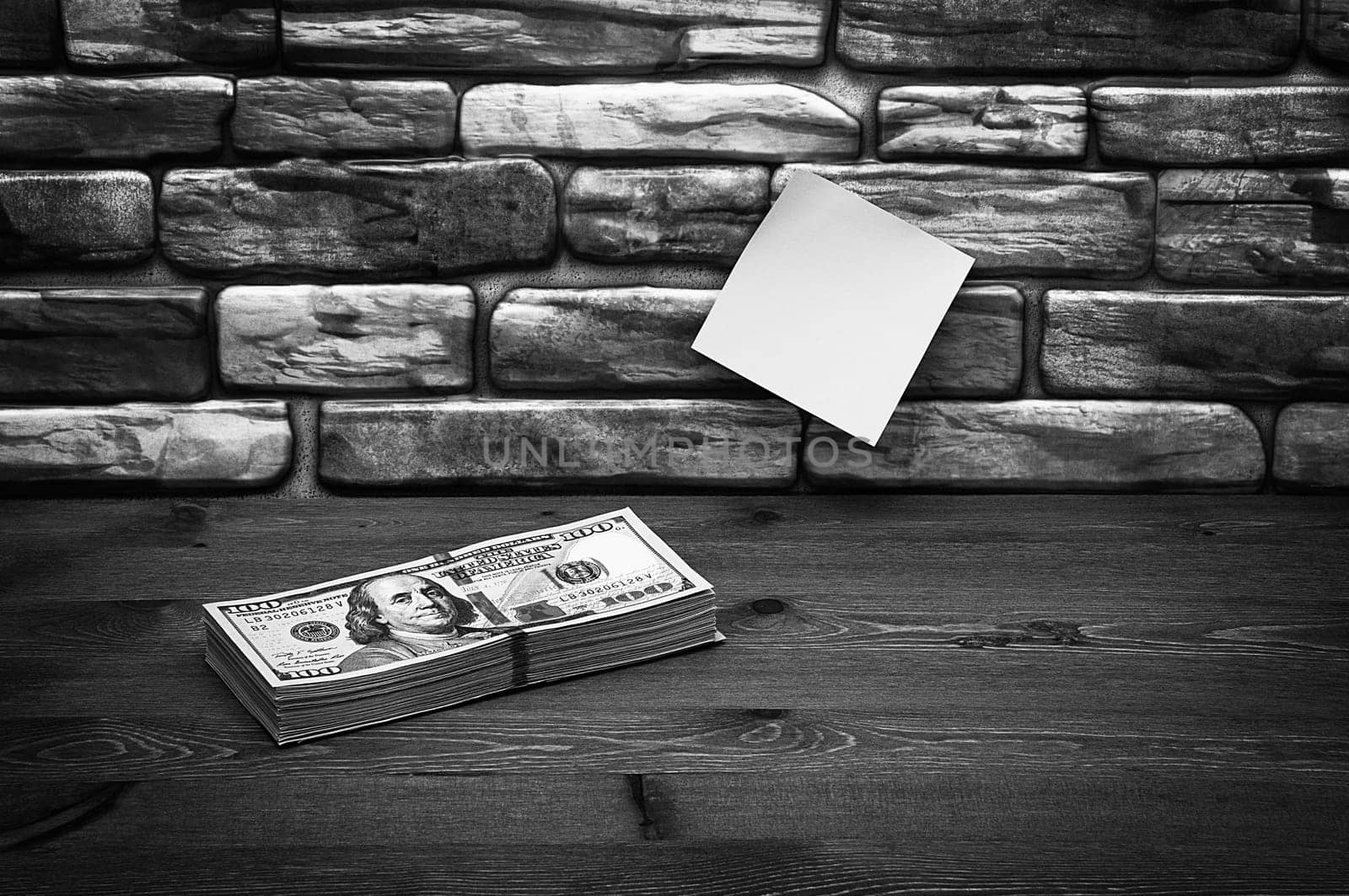 Dollars in one-hundred-dollar bills are stacked on a table near the brick wall. Dollars in a big pile. US dollars. On the brown brick wall there is a place for an inscription, a logo. Black and white style. Layout, template, mockup.