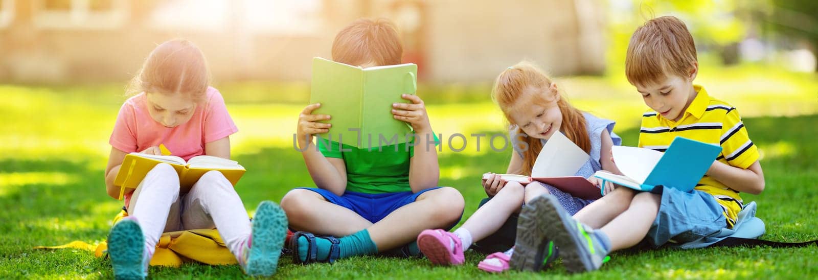 Children with rucksacks sitting in the park on the grass near the school. Pupils with books and backpacks outdoors.