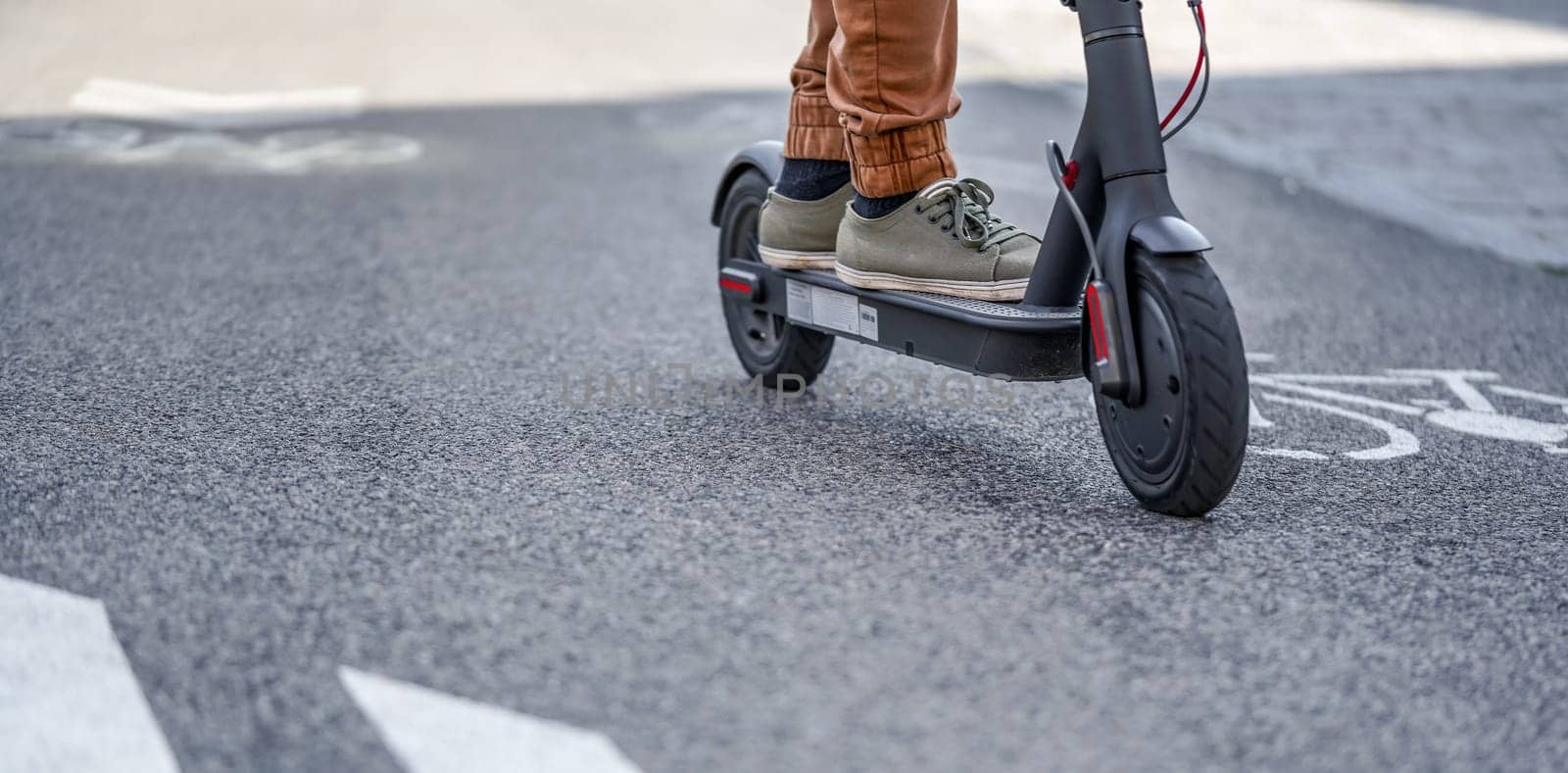 Young man in casual trousers riding an electric scooter detail on his feet and wheel over asphalt road.