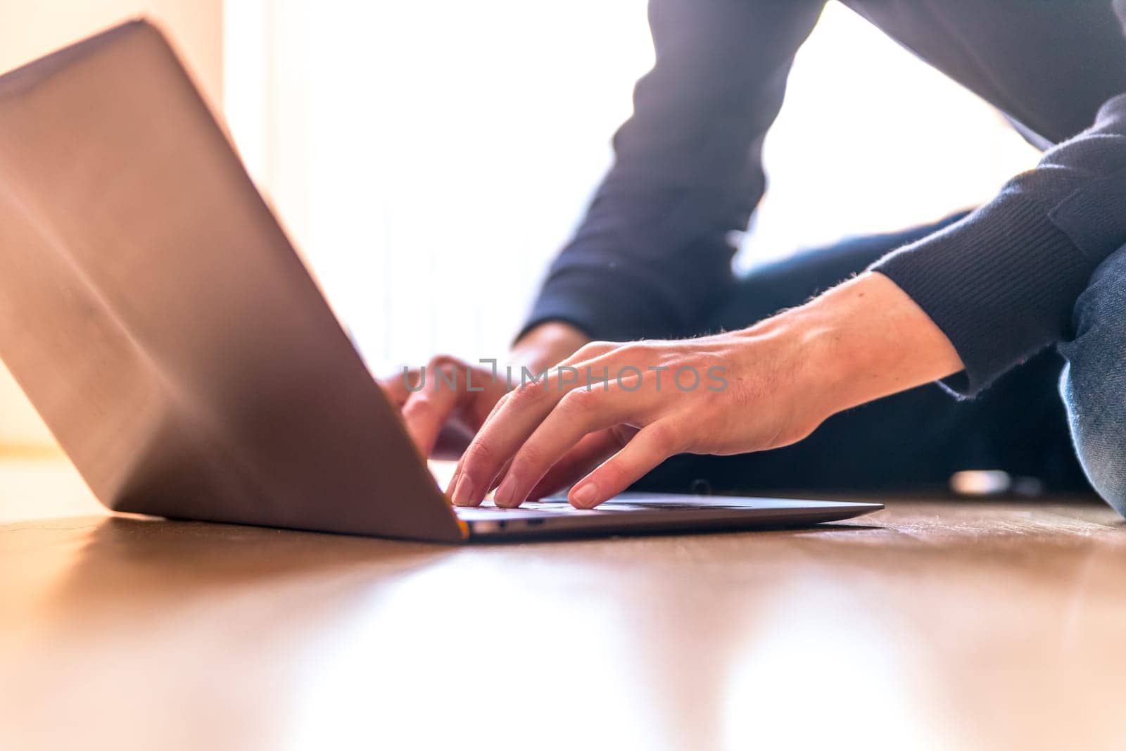 Young man with a laptop is working on the wooden floor, light comes in from the window