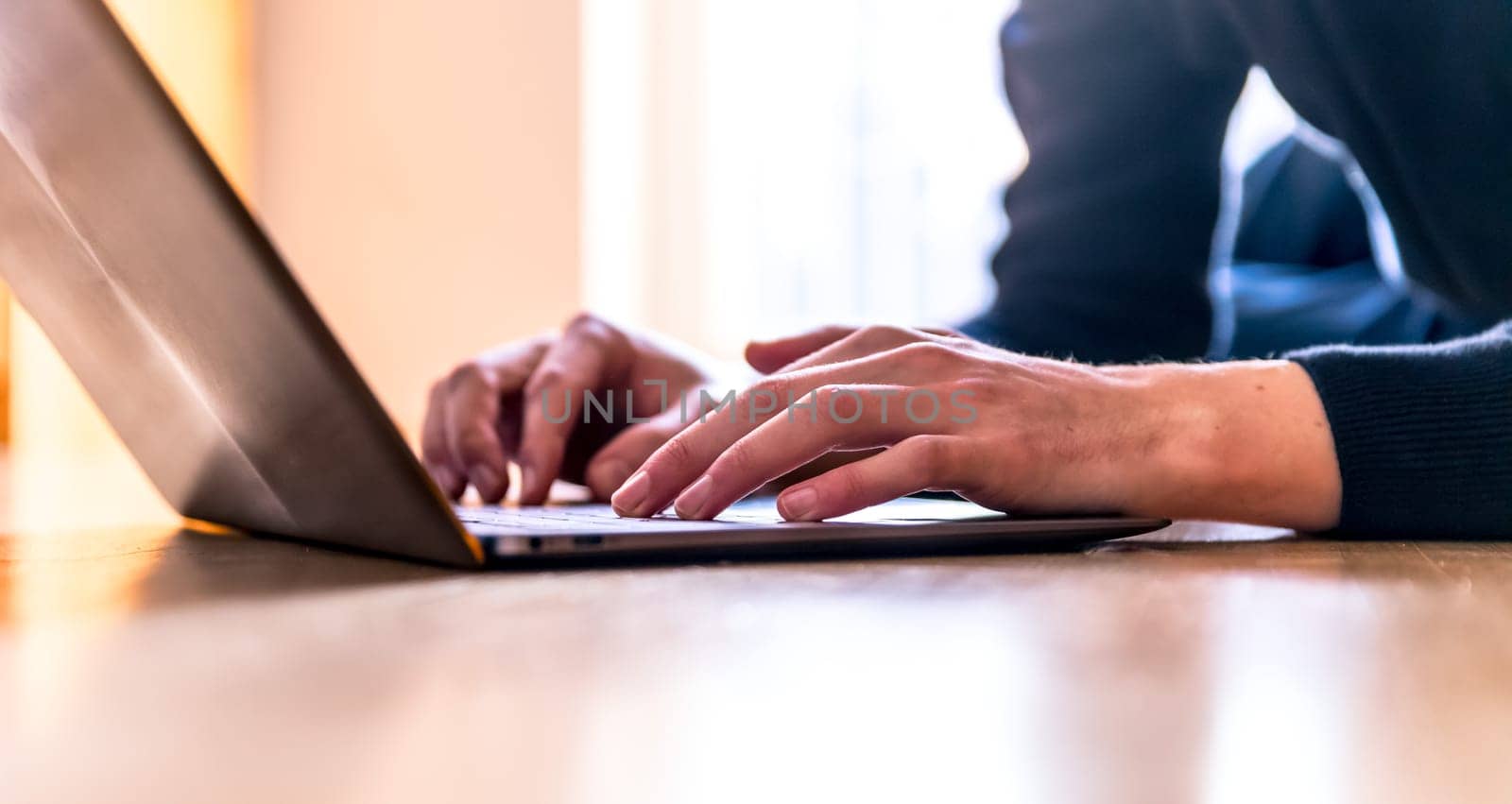 Young man with a laptop is working on the wooden floor, light comes in from the window