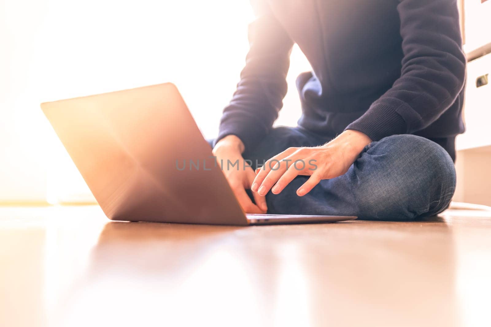 Young man with a laptop is working on the wooden floor, light comes in from the window