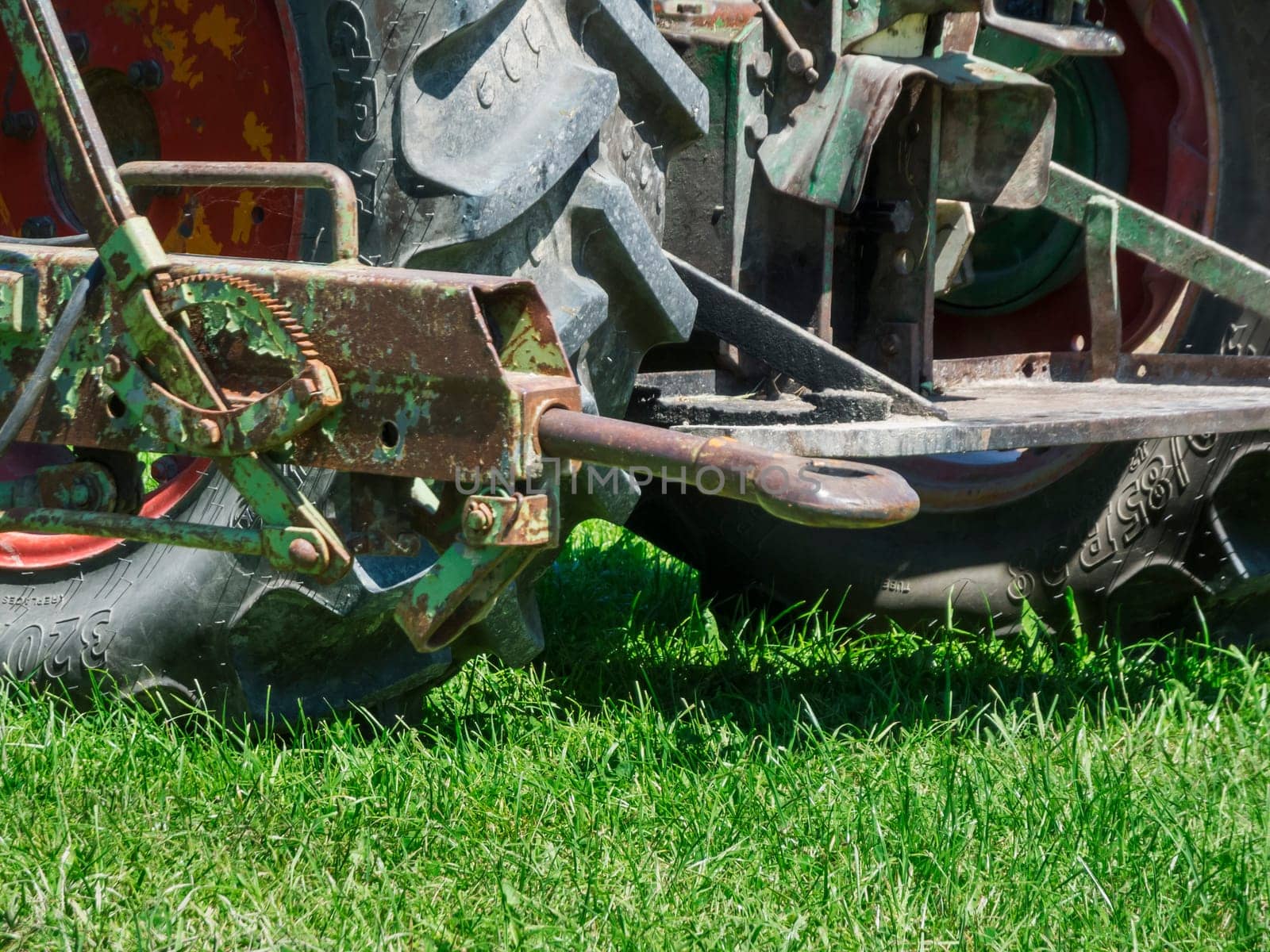Detailed view of an outdated, rusty and bent trailer coupling on a tractor in front of a rough tire tread on a lawn