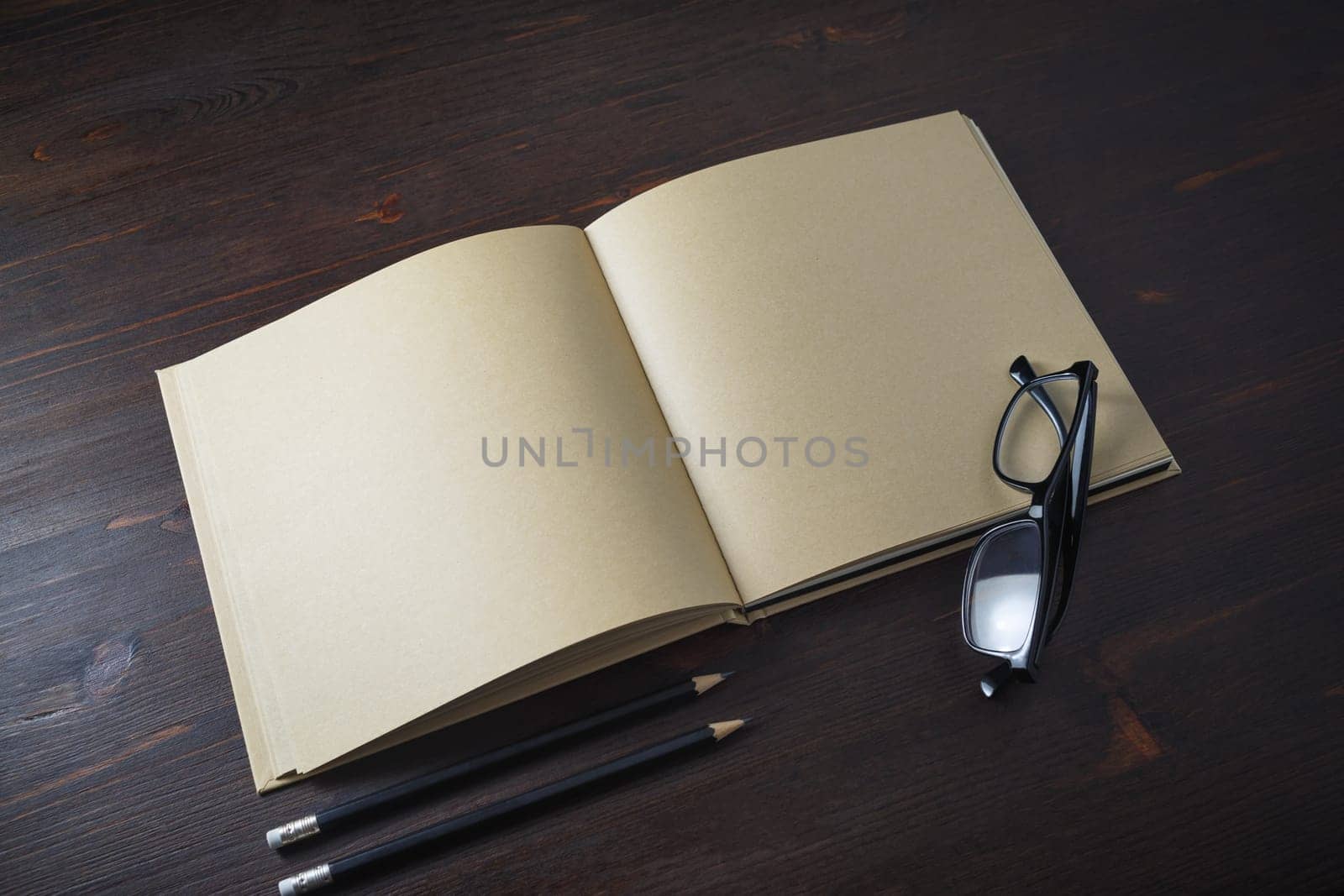 Opened book with blank craft paper pages, pencils and glasses on wooden background.