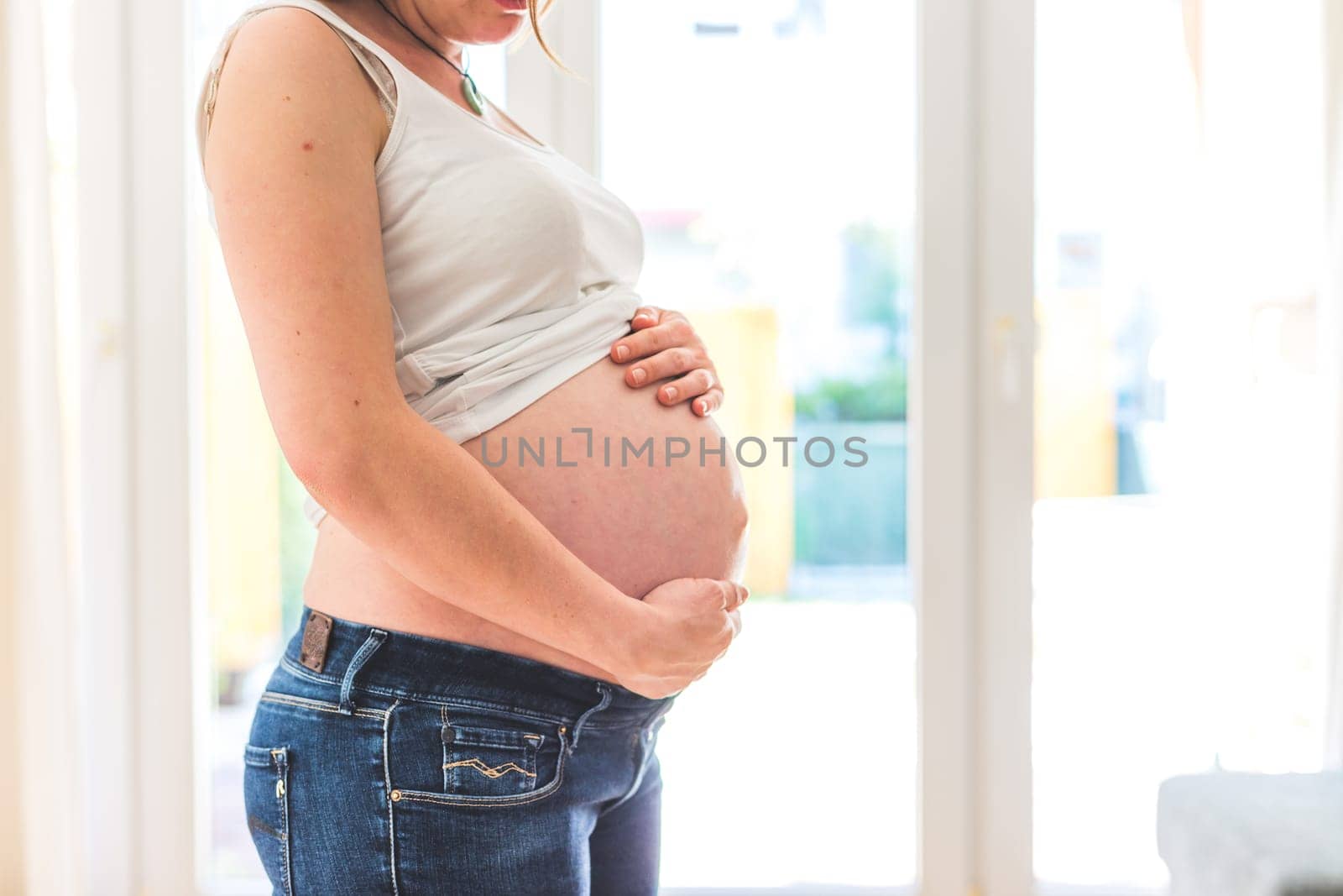 Happy Caucasian pregnant mother touching her naked tummy, blue jeans, close up