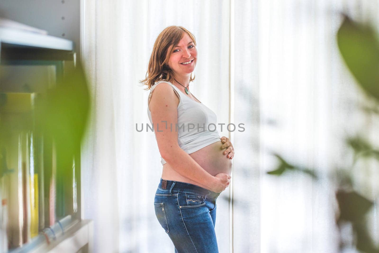 Happy Caucasian pregnant mother touching her naked tummy, blue jeans, close up