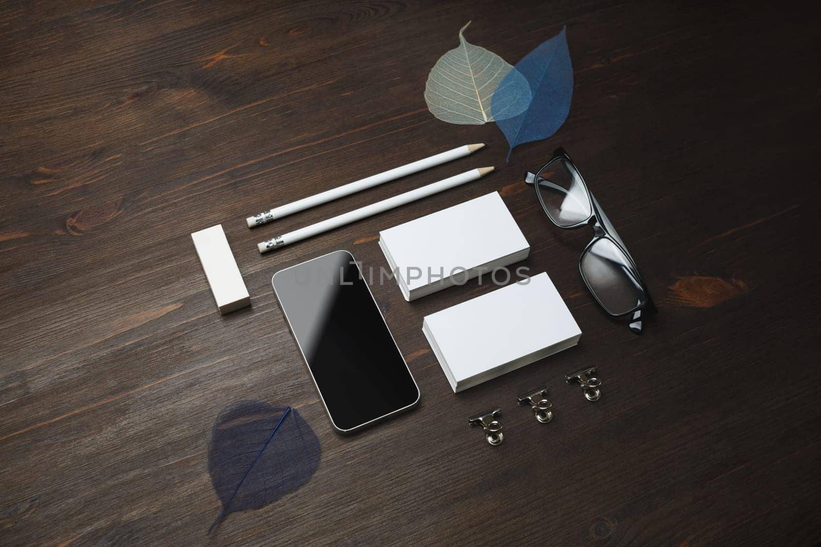 Blank stationery set. Blank business cards, smartphone, pencils, eraser, glasses and leaves on wooden background.