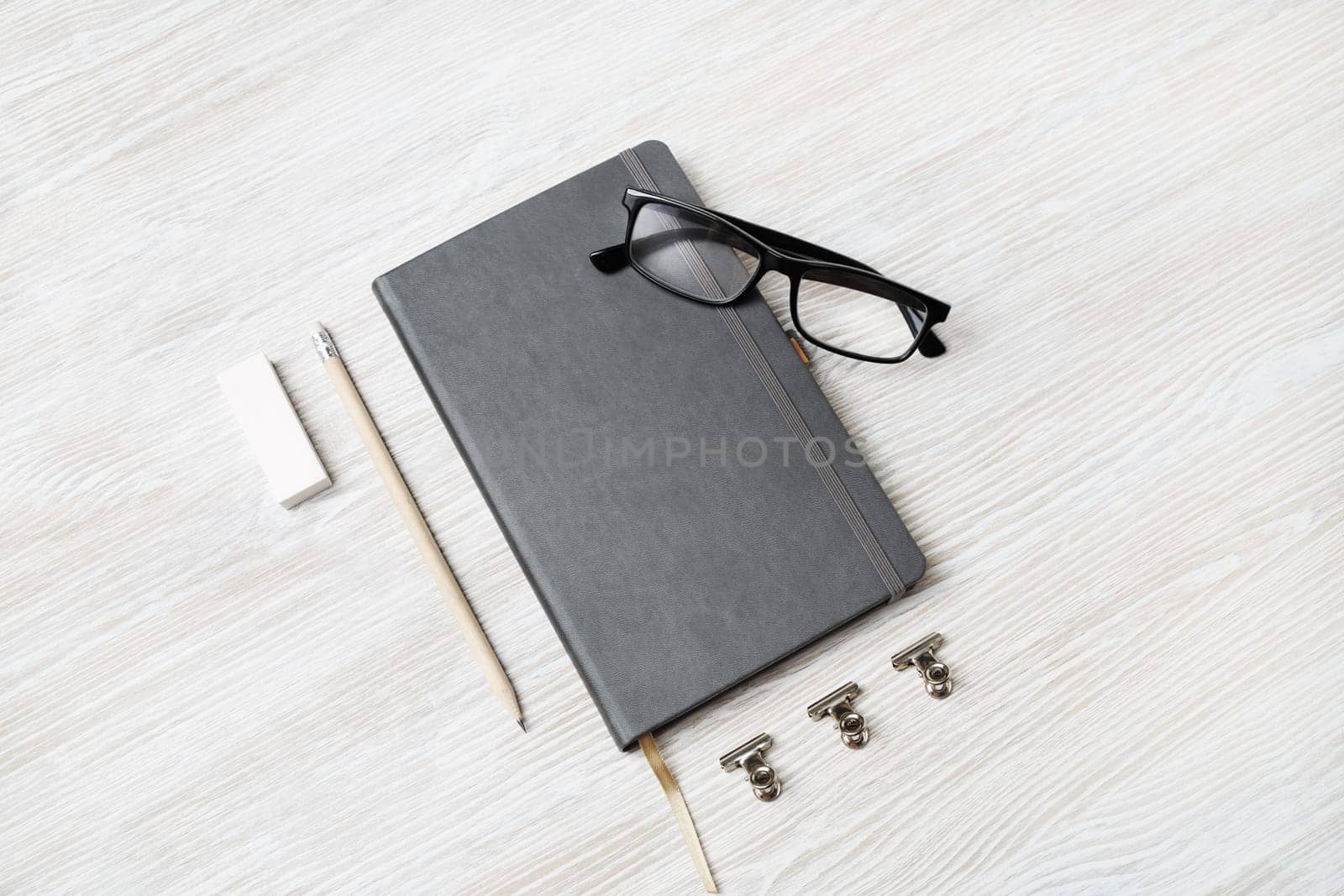Photo of blank closed notebook, glasses, pencil and eraser on light wood table background. Responsive design mockup. Stationery elements.