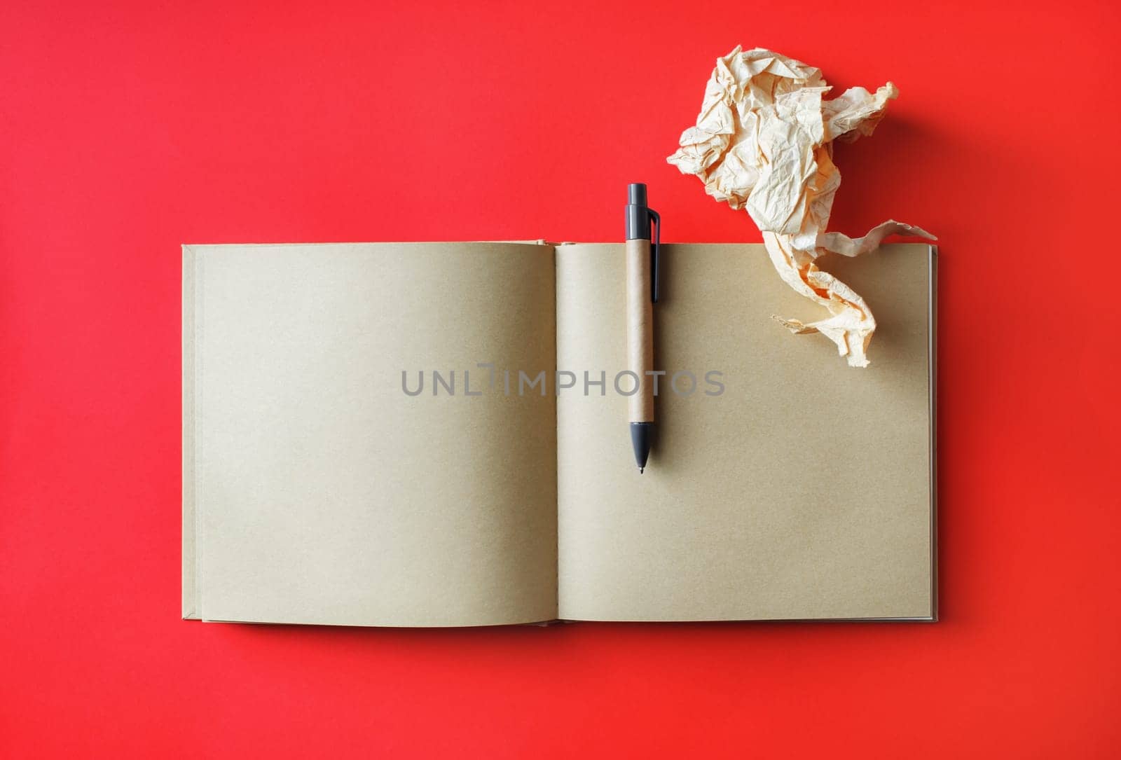 Photo of kraft open book with blank pages, crumpled paper and pen on red paper background. Mockup for placing your design. Top view. Flat lay.