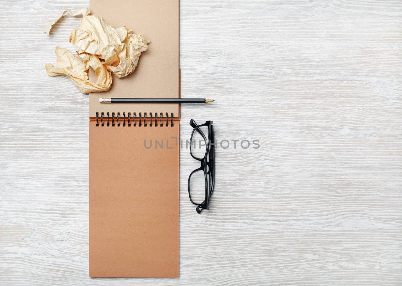 Opened notebook with blank craft paper pages, glasses, pencil and crumpled paper on light wood table background. Flat lay.