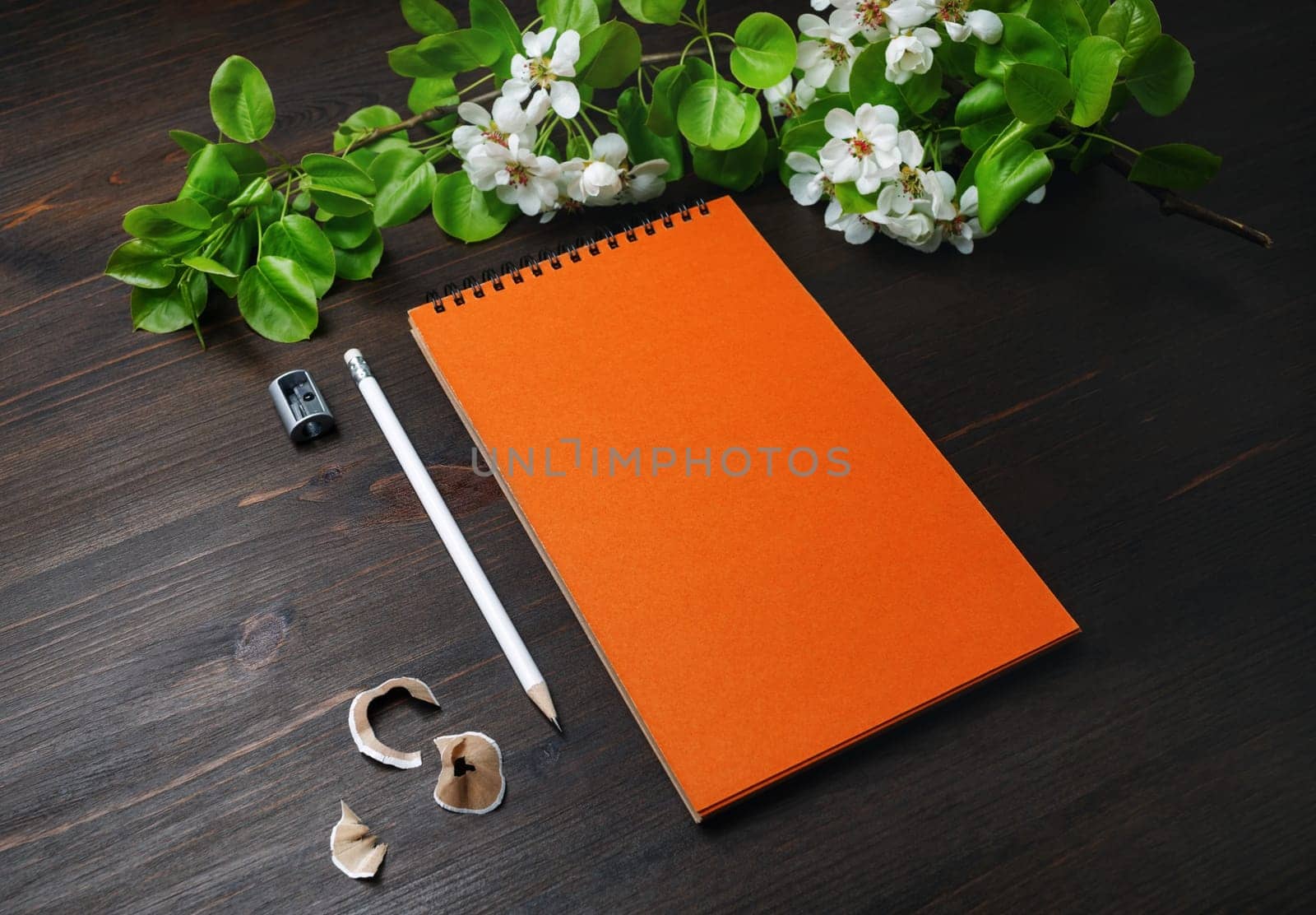 Photo of blank stationery template. Orange sketchbook, pencil, sharpener and spring flowers on wood table background.