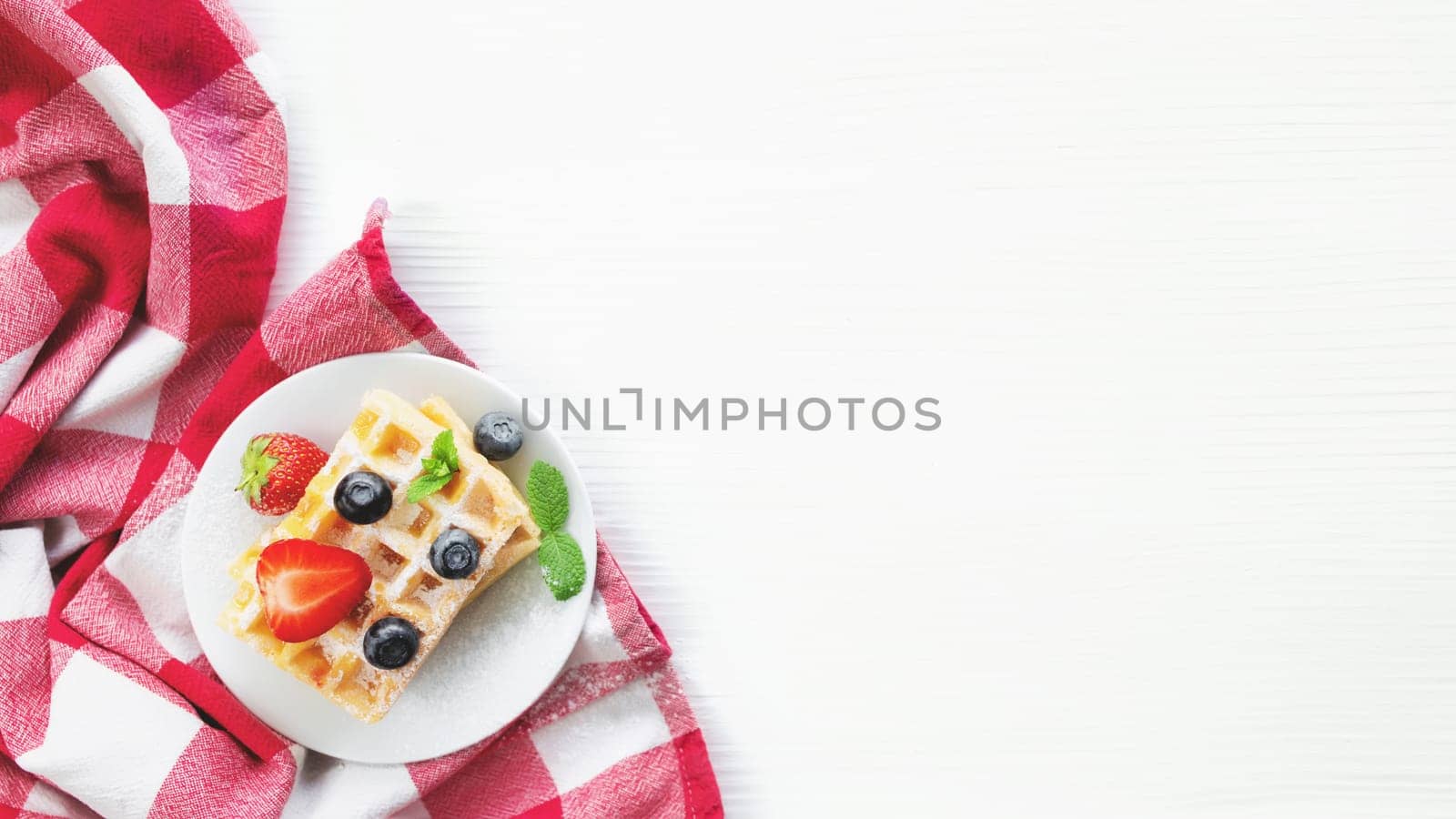White plate on red kitchen towel with stack homemade Belgian waffles, blueberry, strawberry cuts, mint leaf, sugar powder on gray background