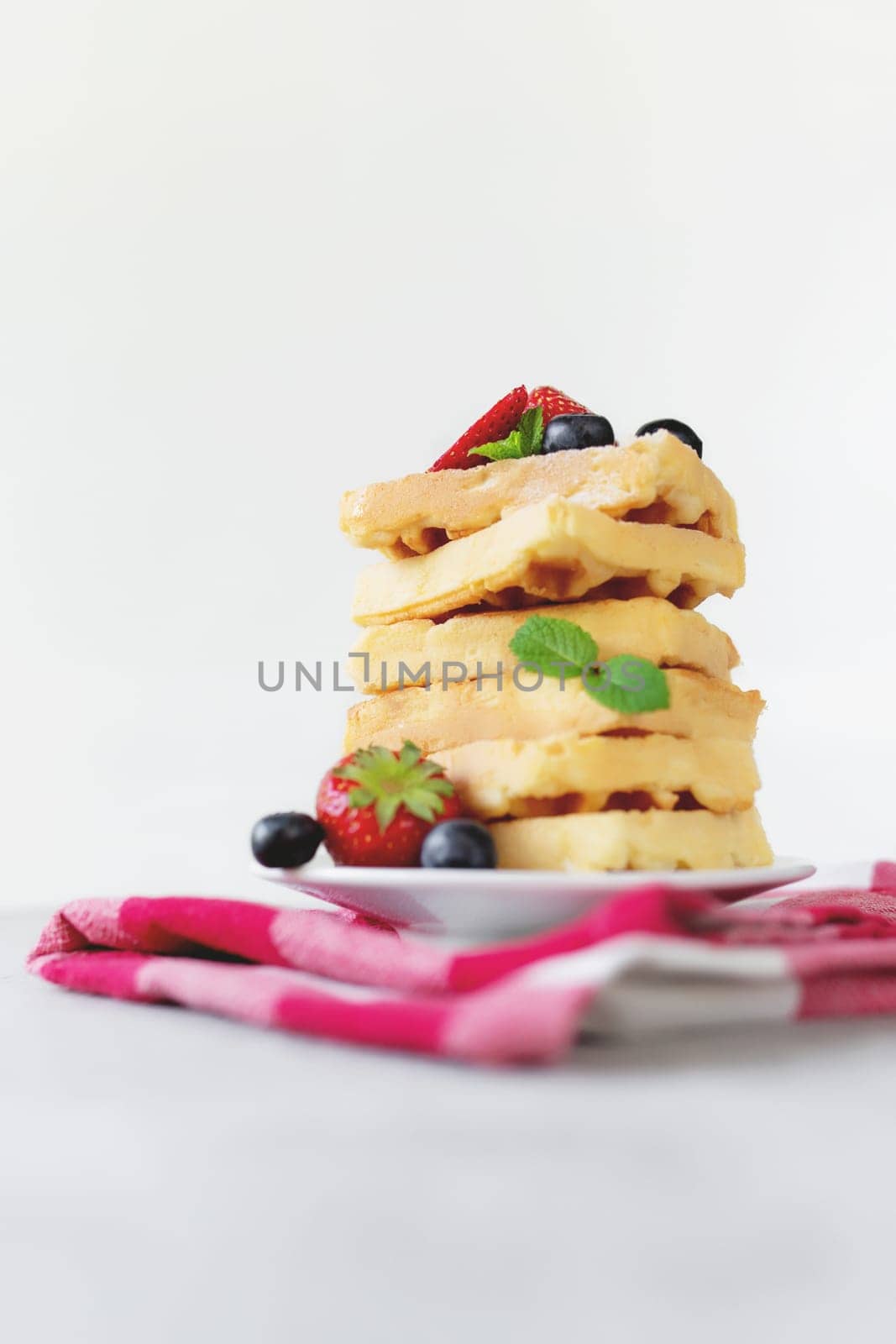 White plate on red kitchen towel with stack homemade Belgian waffles, blueberry, strawberry cuts, mint leaf, sugar powder on gray background