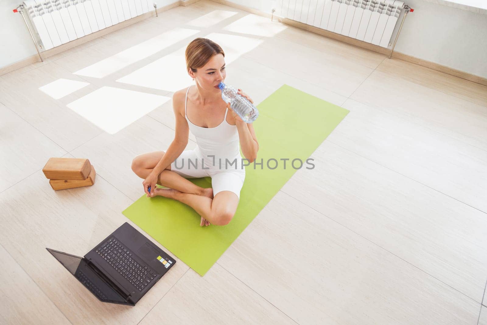 Woman sitting on mat in sportswear, drinks water and doing sport at home with laptop online