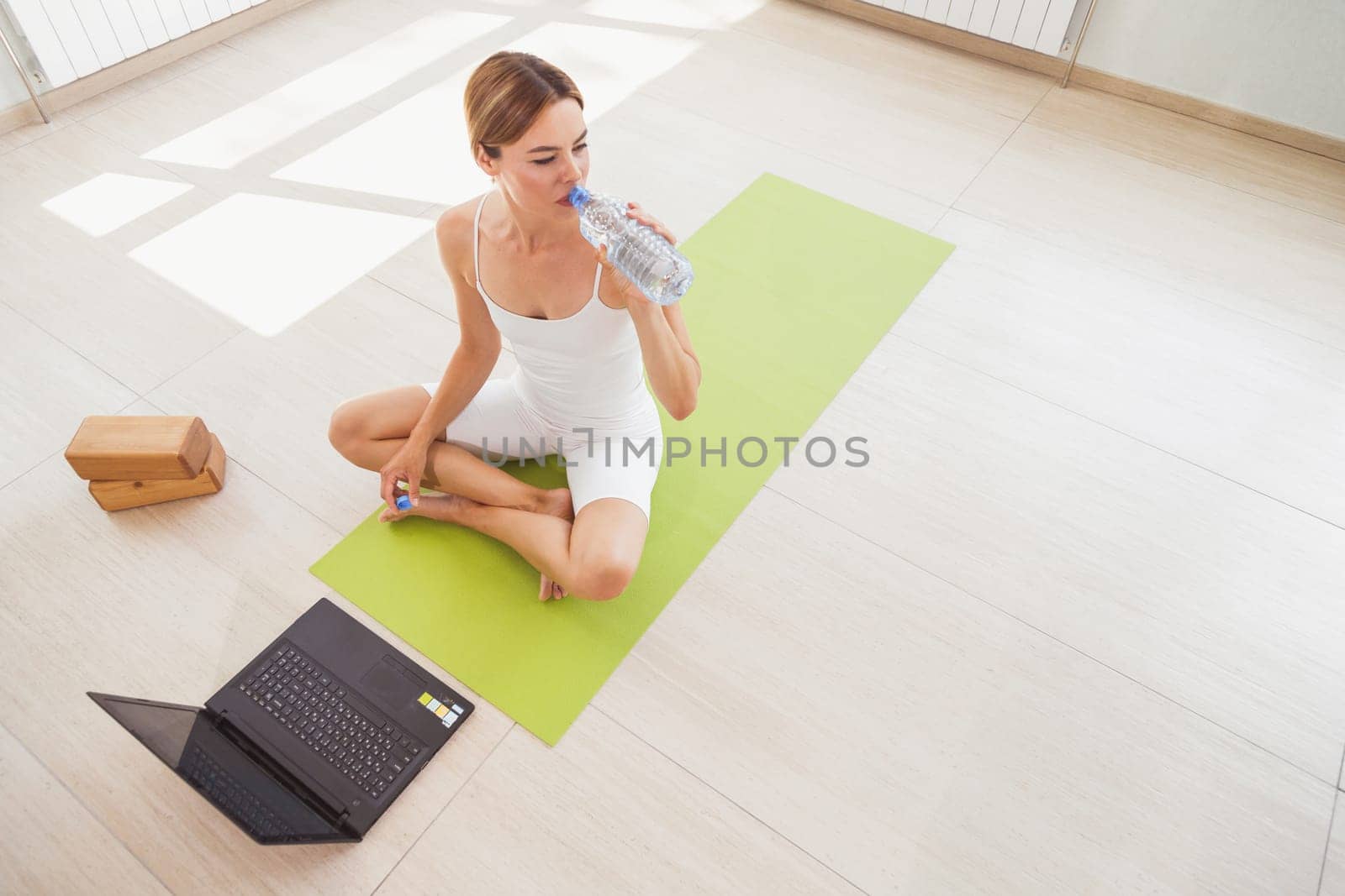 Woman sitting on mat in sportswear, drinks water and doing sport at home with laptop online