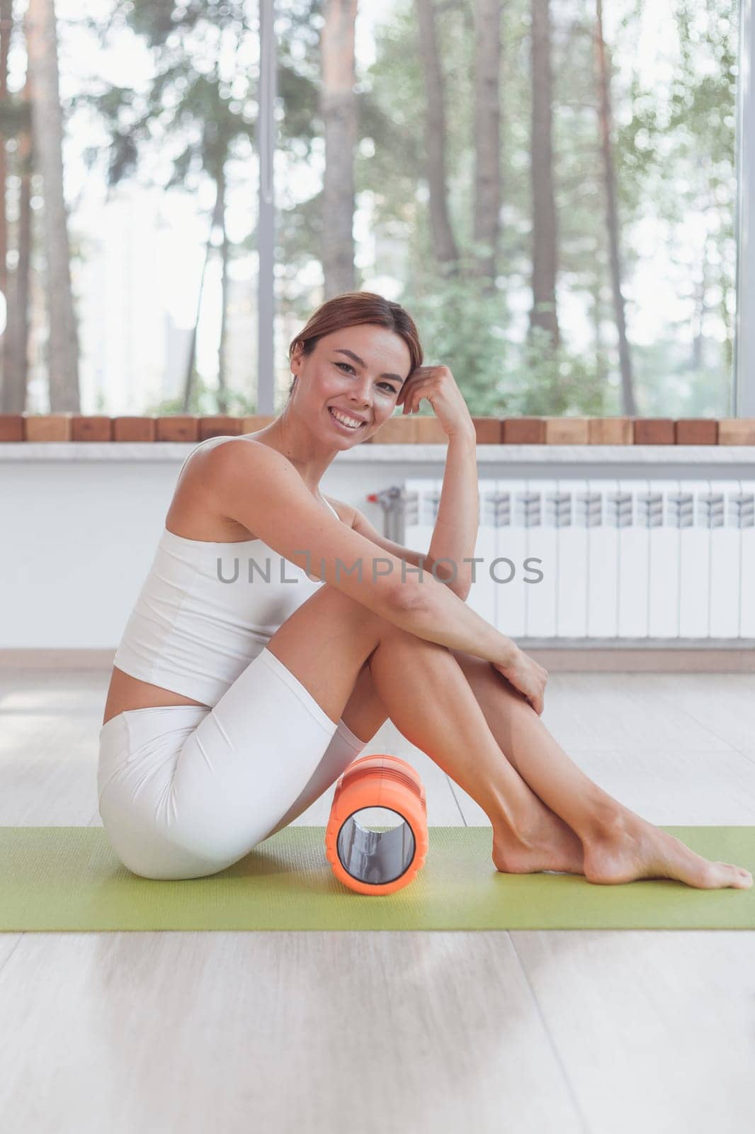 Sporty woman with foam roller sitting on mat in sport hall