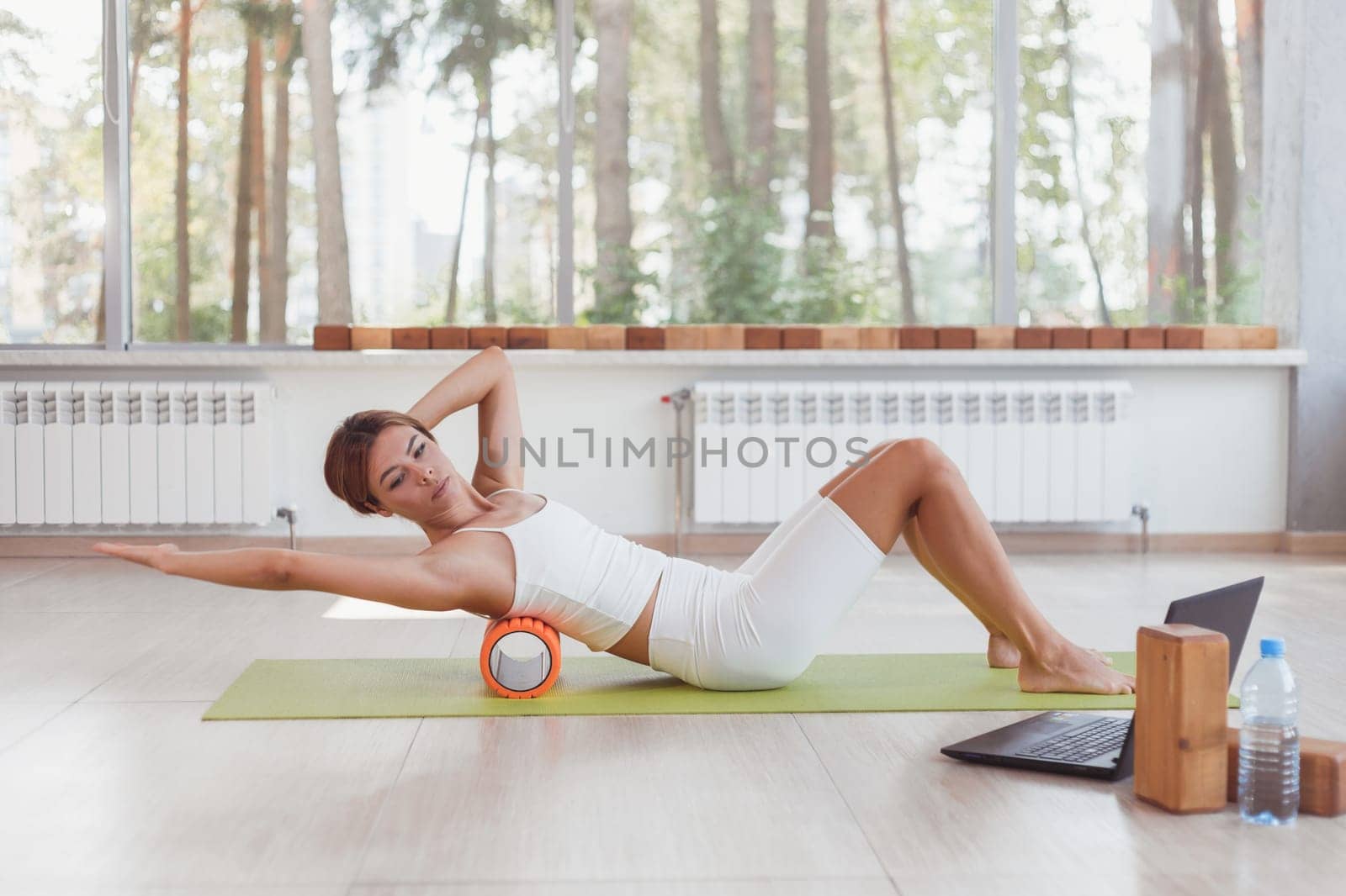 Sporty woman doing online exercises with foam roller under the back. Workout online with laptop. Instructor online