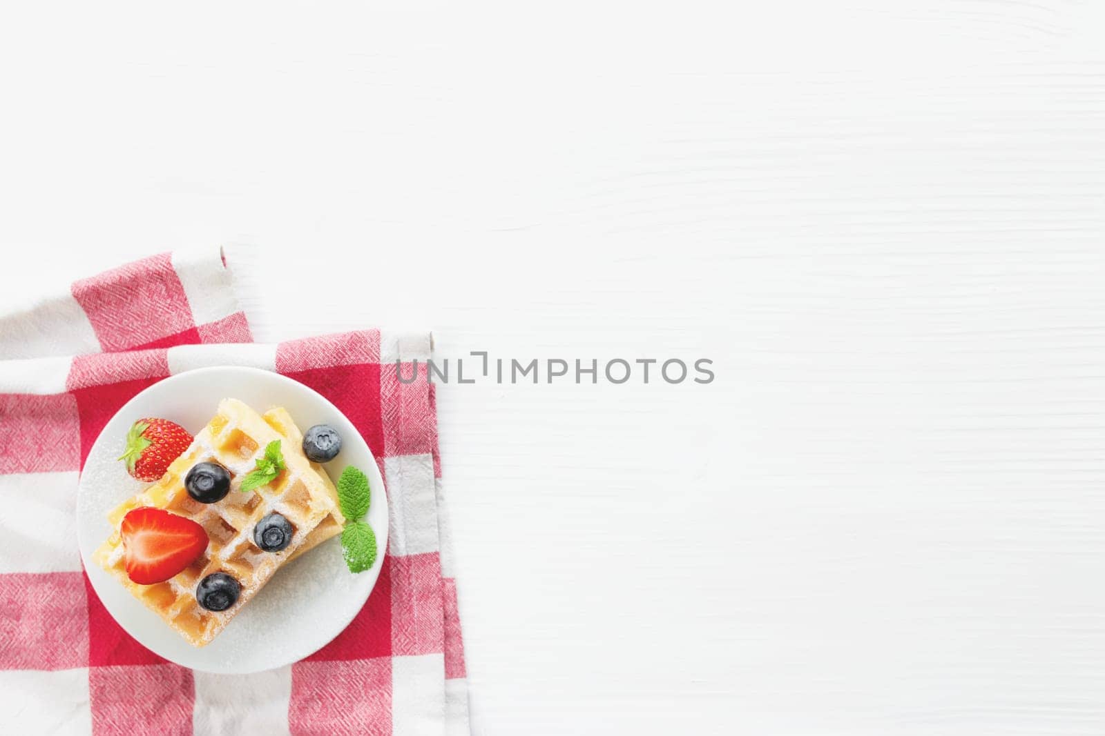 White plate on red kitchen towel with stack homemade Belgian waffles, blueberry, strawberry cuts, mint leaf, sugar powder on gray background