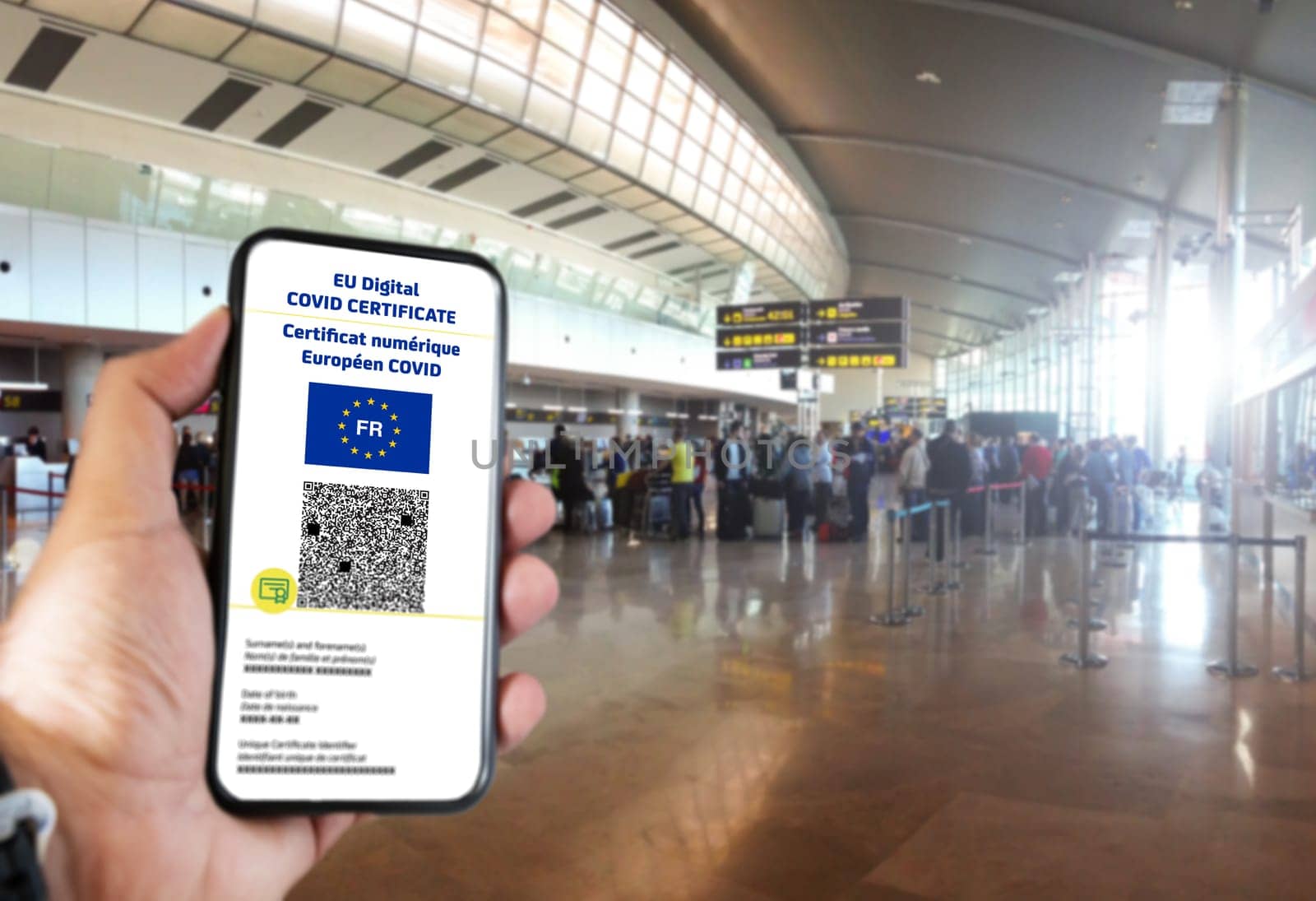 French EU Digital COVID Certificate with the QR code on the screen of a mobile held by a hand with blurred airport in the background. Translation from french "European COVID digital certificate"""